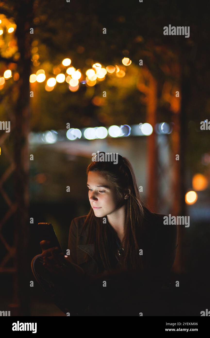 Eine lächelnde junge Frau, die nachts ein Smartphone benutzt. Sie sitzt im Garten mit einem Haus im Hintergrund Stockfoto