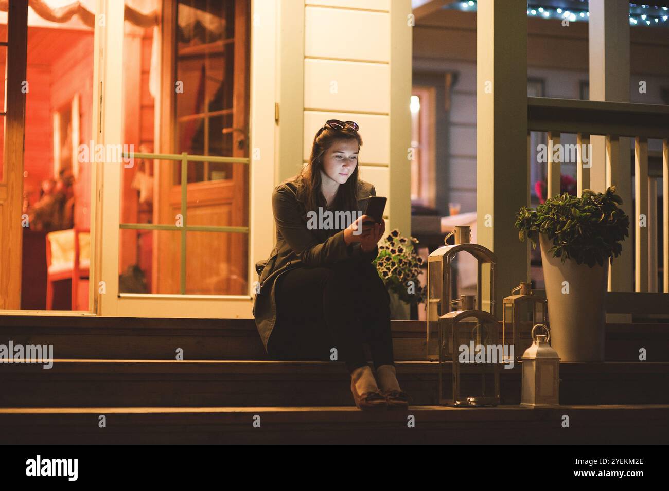 Eine lächelnde junge Frau, die nachts ein Smartphone benutzt. Sie sitzt im Garten mit einem Haus im Hintergrund Stockfoto