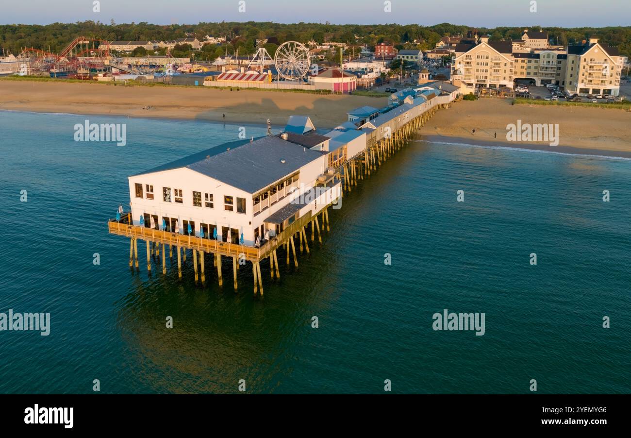 Der Old Orchard Beach Pier erstreckt sich bis ins Meer, mit Gebäuden auf Stelzen und erstreckt sich von einem Sandstrand, der von einem Vergnügungspark gesäumt wird. Ferris mit Stockfoto
