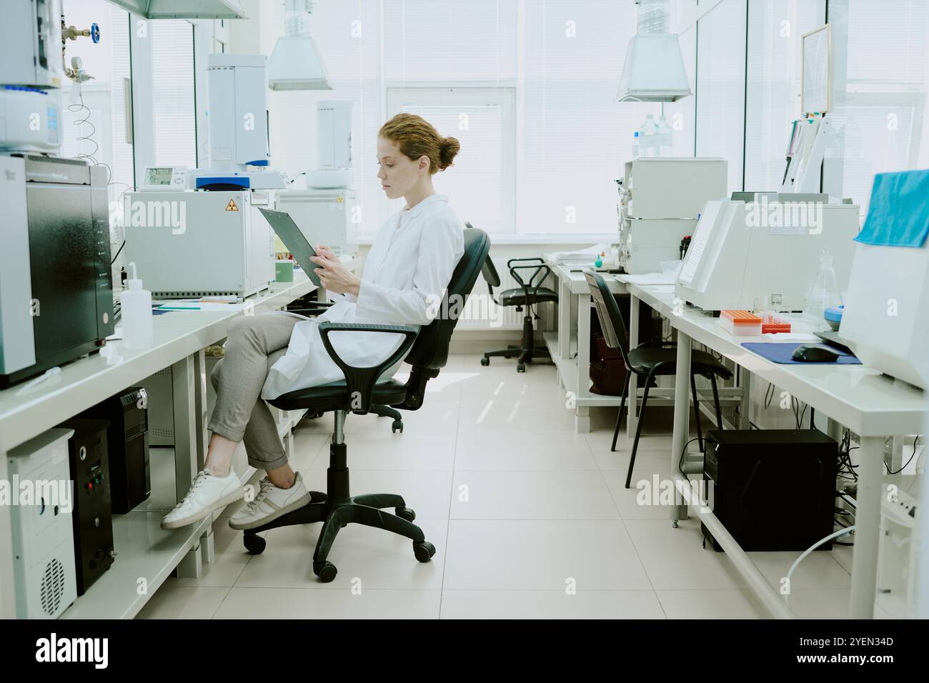 Wissenschaftlerinnen, die Laborkittel tragen, sitzen auf einem Stuhl in einem modernen Labor, umgeben von Geräten, mit einem digitalen Tablet, das sich auf die Arbeitsumgebung konzentriert Stockfoto