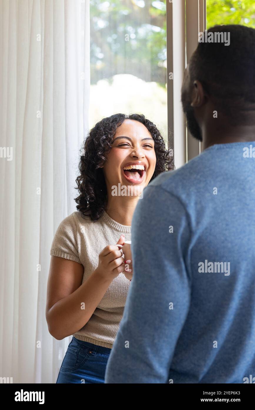 Lachende Frau, die eine Kaffeetasse hält und sich mit dem Partner zu Hause unterhalten kann. Glück, multirassisches Paar, Lebensstil, Bindung, Morgen Stockfoto