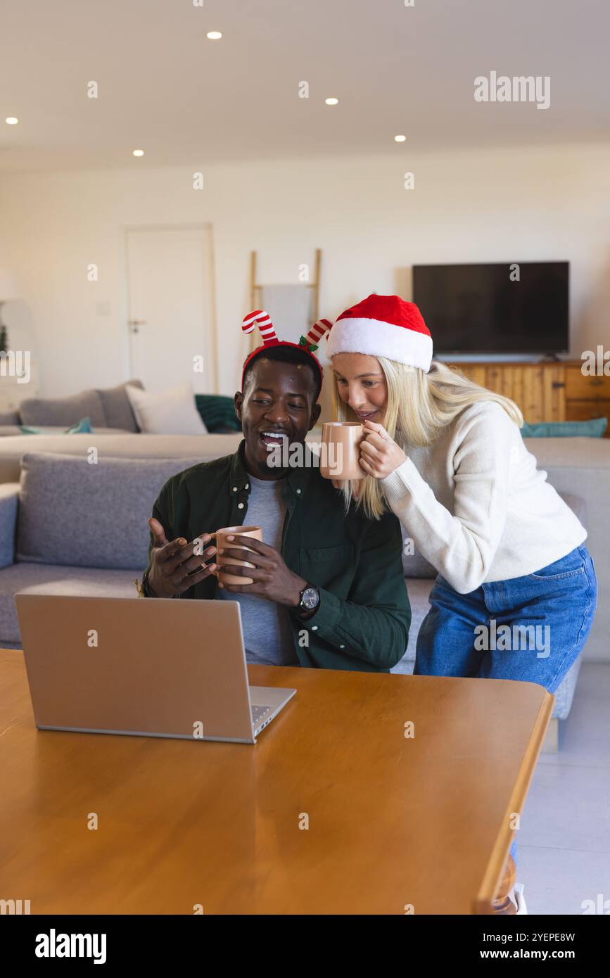 Weihnachtszeit, multirassisches Paar genießt Videoanrufe zu Hause mit festlichen Hüten und Tassen. Feiern, Vielfalt, Kommunikation, Glück, virtuell, Stockfoto