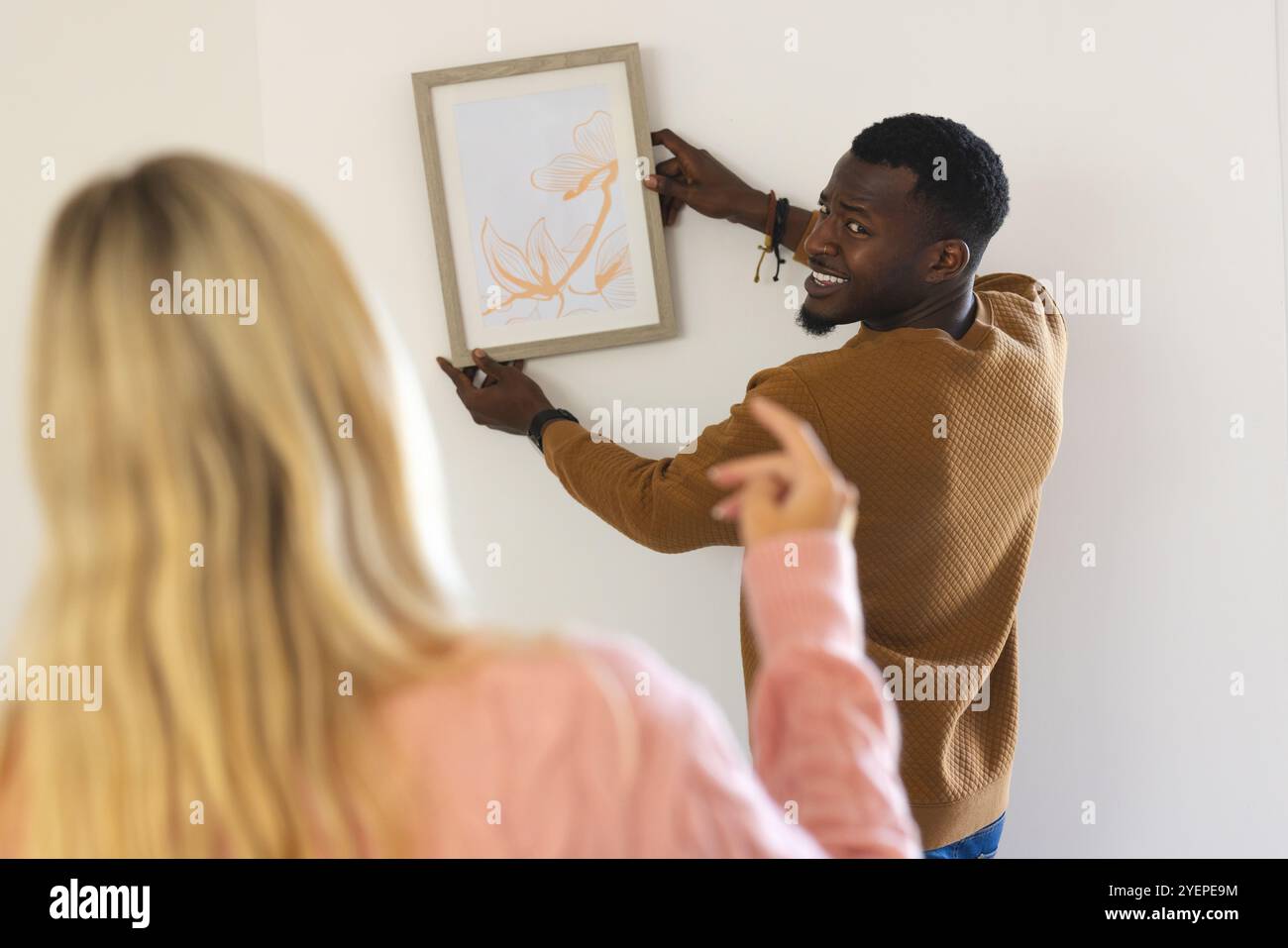 Ein afroamerikanischer Mann hängt ein Bild, während die Frau zu Hause Wegbeschreibungen gibt. Dekoration, Teamarbeit, Familie, Kreativität, Kunst, Wohnraum Stockfoto