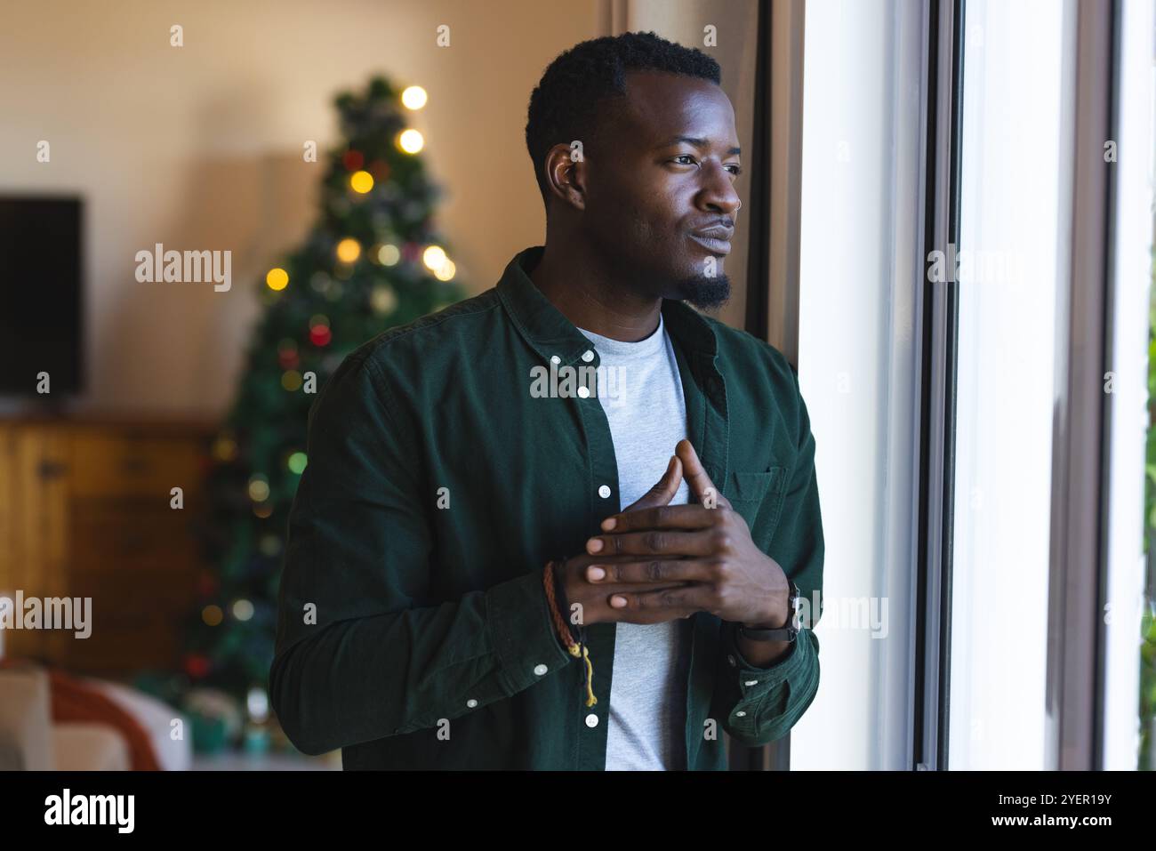 Am Fenster steht ein afroamerikanischer Mann, der über die Weihnachtszeit mit einem Weihnachtsbaum in der Nähe nachdenkt. Feiertage, Festtage, Dekoration, Kontemplation, Feiern Stockfoto