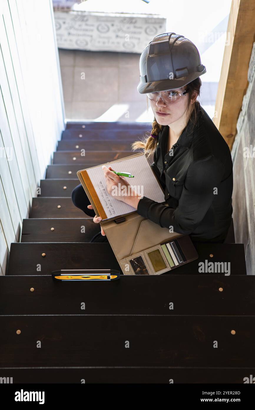 Die Bauinspektorin sitzt auf der Treppe und macht Notizen auf ihrem Klemmbrett. Während der vor dem Kauf durchgeführten Inspektion der Innentreppen des Hauses. Vertikal Stockfoto