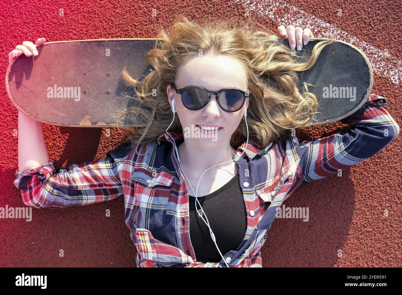 Millennial Hippie Frau mit Sonnenbrille und Kopfhörer auf einem Skateboard auf der Straße liegend. Skater Mädchen auf einem Longboard. Coole Skateboarderin bei s Stockfoto