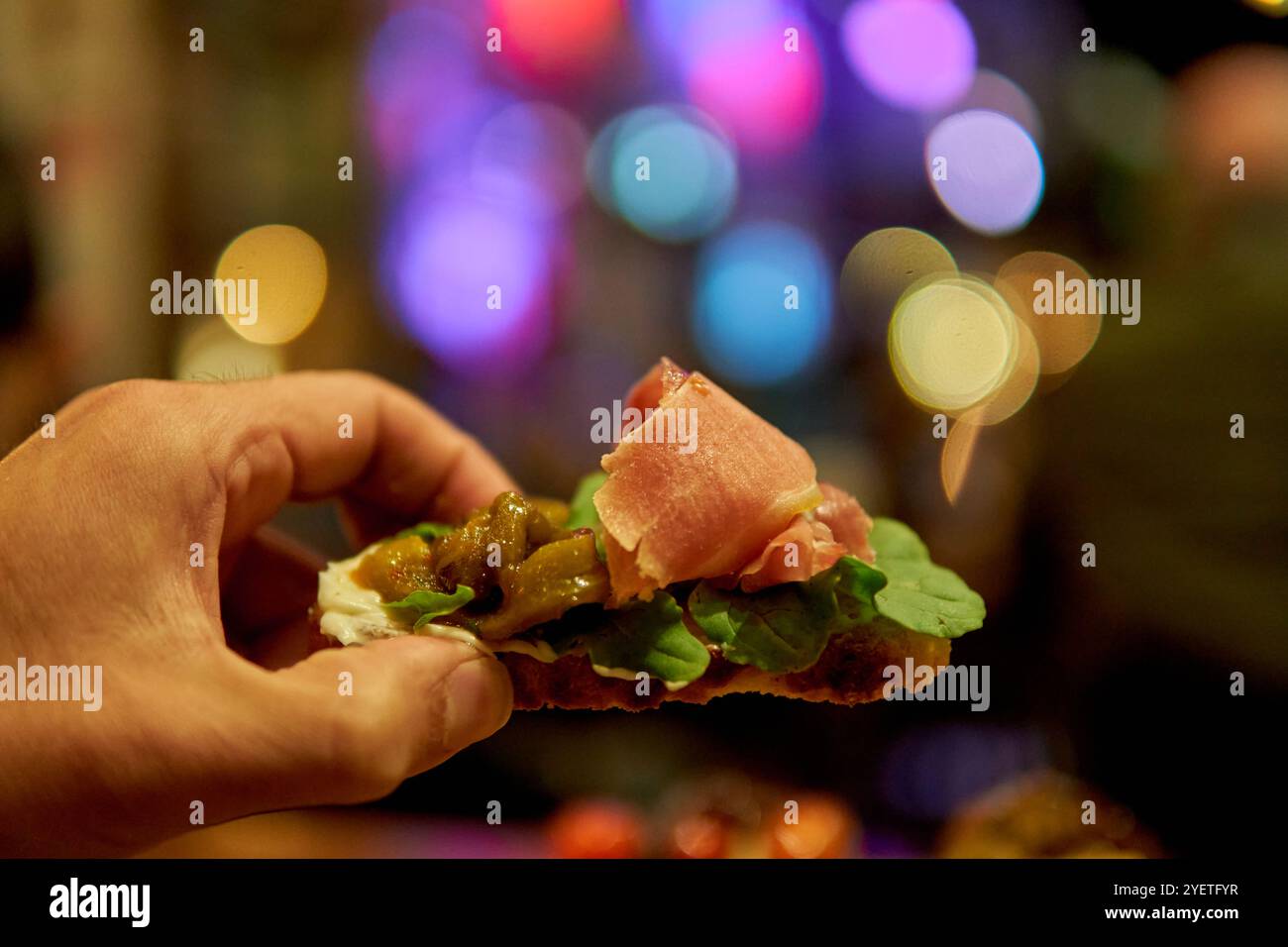 Nahaufnahme einer männlichen Hand, die eine Bruschetta aus Rucola, Schinken und eingelegter Aubergine vor einem Hintergrund aus unscharfen farbigen Lichtern hält Stockfoto