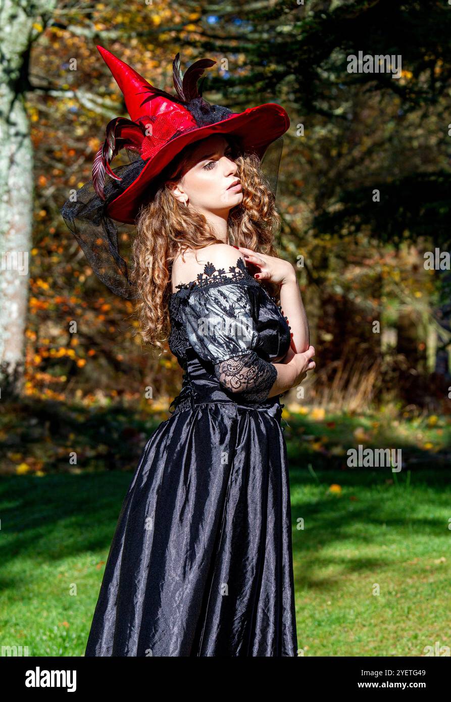 Eine Hexe in einem langen schwarzen Kleid und einem Hexenhut im Park draußen in der rauen Herbstsonne in Schottland Stockfoto