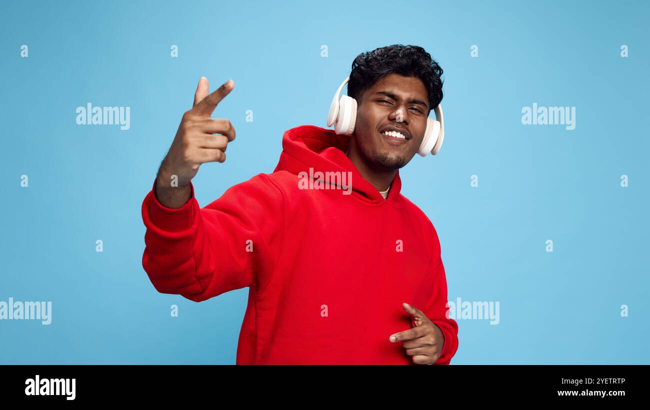 Porträt eines jungen Indianers in rotem übergroßem Hoodie, der energische Musik in Kopfhörern hört und vor blauem Studiohintergrund singt Stockfoto