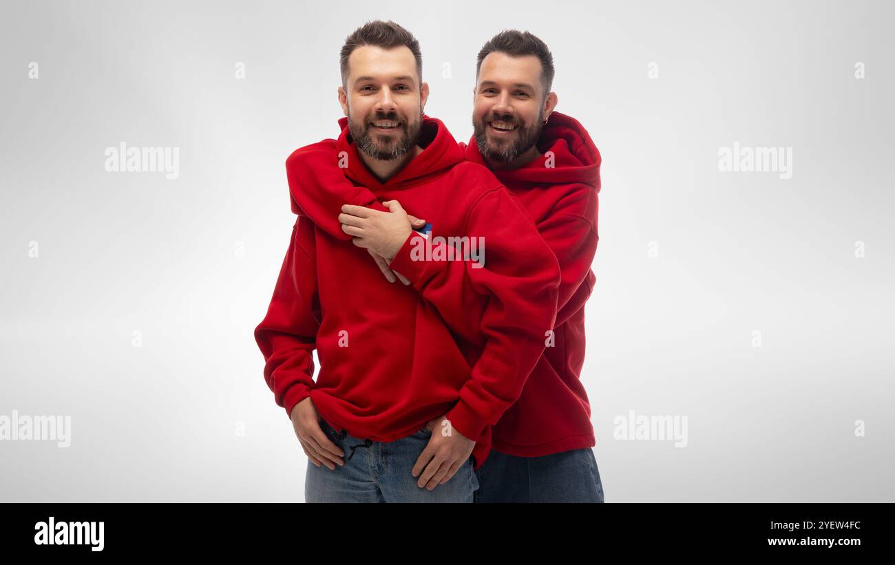 Zwei junge Männer, Geschwister, die zu zwanglosen Outfits gekleidet sind, umarmen sich und blicken mit fröhlichem Lächeln in die Kamera, um Freude über das Treffen mit weißem Studio auszudrücken Stockfoto