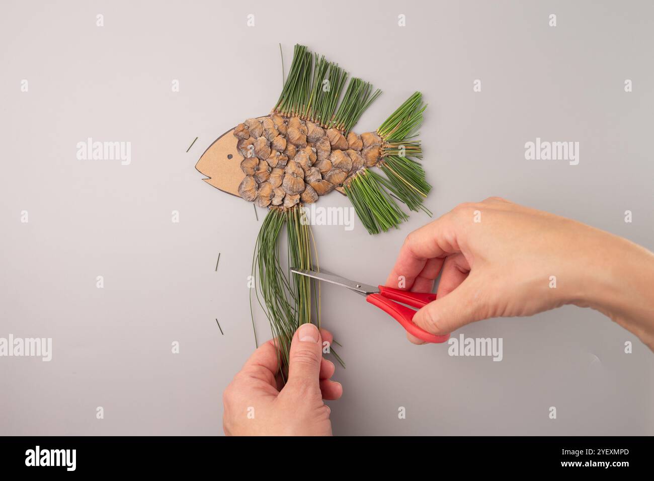 Person schneidet fischförmiges Handwerk aus Tannenzapfen und Tannennadeln mit roter Schere auf grauem Hintergrund. Praktisches DIY-Projektkonzept. Flach liegend Co Stockfoto