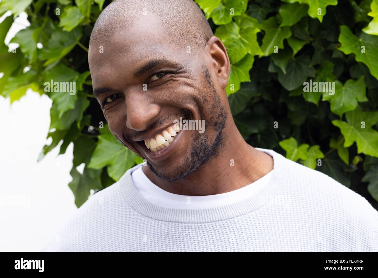 Lächelnder afroamerikanischer Mann in weißem Pullover, der draußen mit grünem Blatthintergrund steht Stockfoto