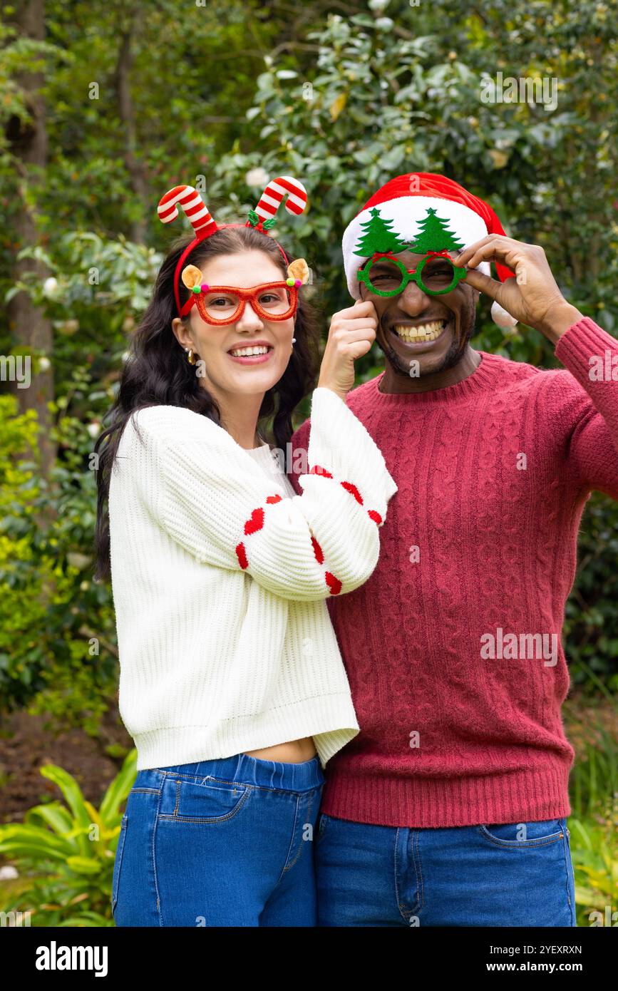 Glückliches multirassisches Paar, das festliche Brillen und Pullover trägt und Weihnachten im Freien feiert Stockfoto