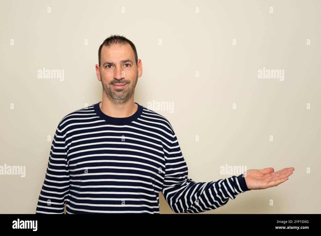 Ein bärtiger hispanischer Mann in seinen 40ern, der einen gestreiften Pullover trug, der seine Hand zur Seite streckte, in einer Geste des Darbietens oder Ausdehnens, indem er ihn in der Hand hielt Stockfoto