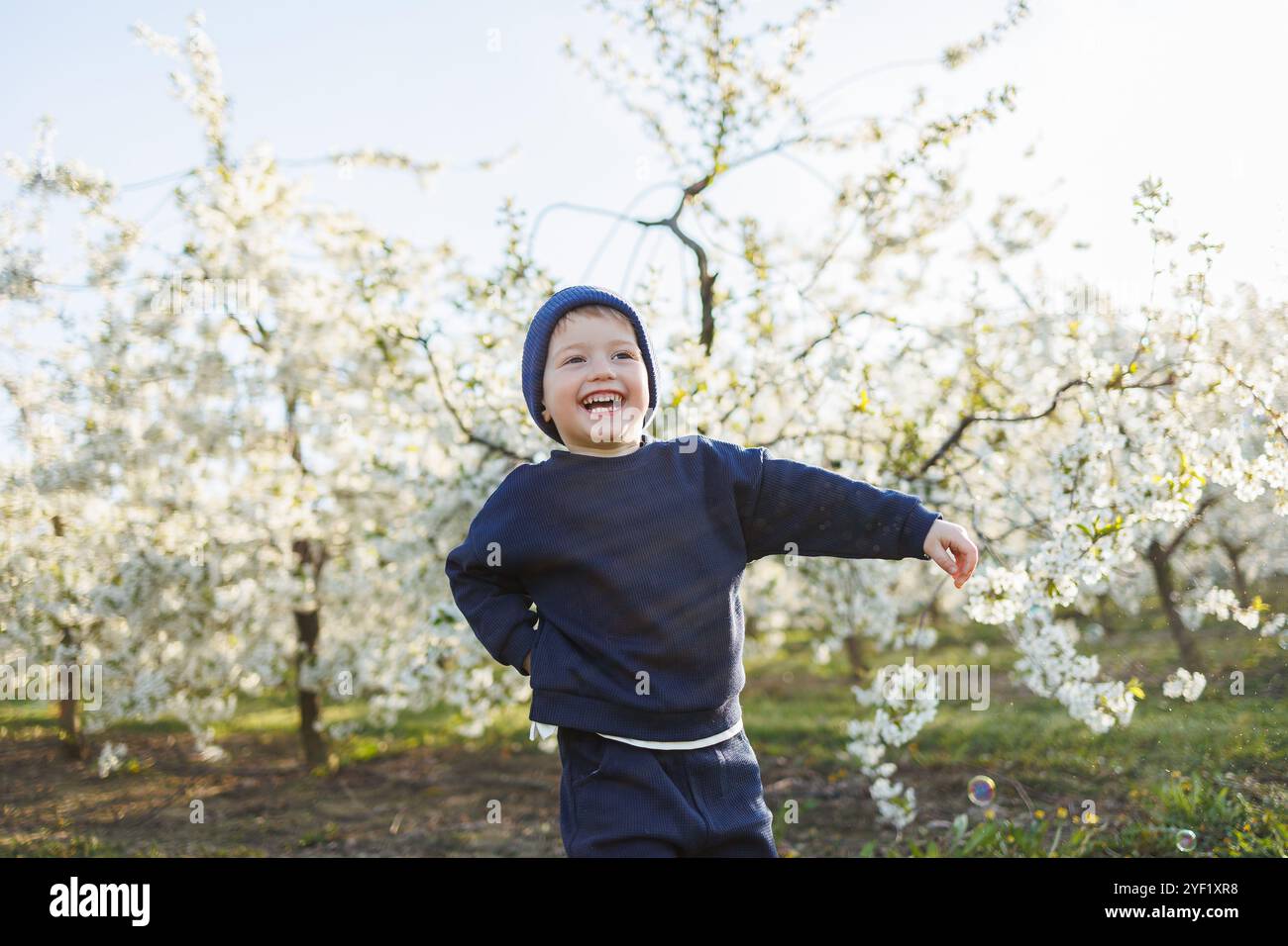 Fröhlicher Junge, 3 Jahre alt, auf einem Spaziergang in einem blühenden Garten. Ein kleiner Junge, 3 Jahre alt, in Pullover und Hut, läuft durch einen blühenden Garten. Kleidung für Stockfoto