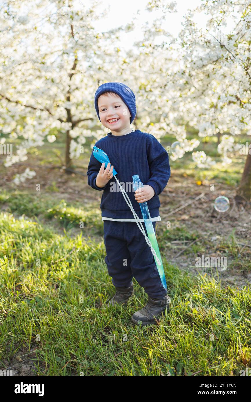 Ein dreijähriger Junge läuft mit Seifenblasen durch einen blühenden Garten. Fröhliches emotionales Kind läuft im Park. Seifenleisten für Kinder Stockfoto