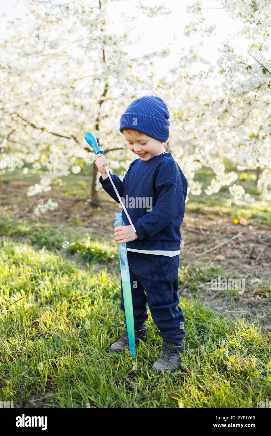 Ein dreijähriger Junge läuft mit Seifenblasen durch einen blühenden Garten. Fröhliches emotionales Kind läuft im Park. Seifenleisten für Kinder Stockfoto