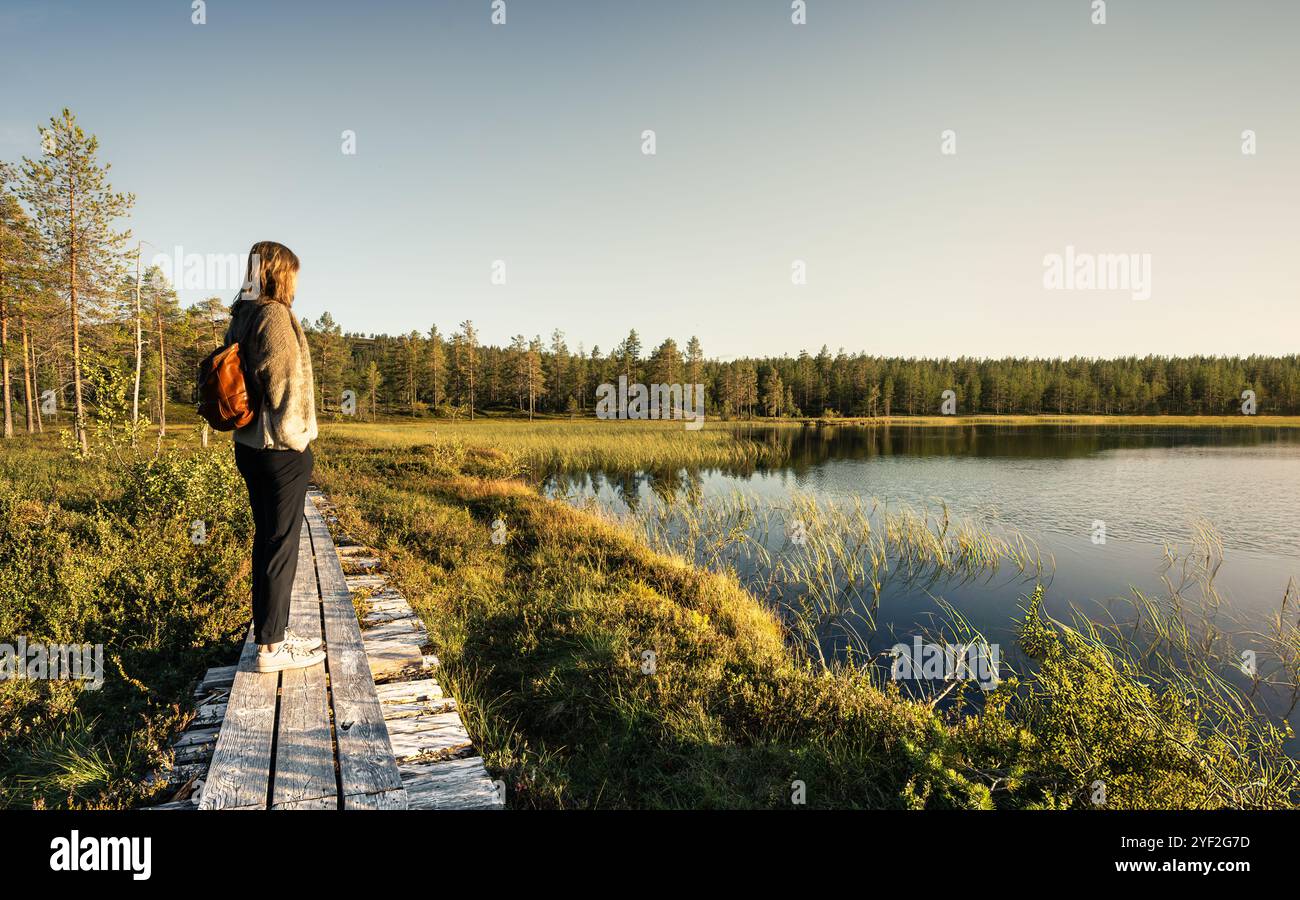 Frau, die auf einem Holzsteg steht und einen See überblickt, der vom Wald umgeben ist, während die Abendsonne ein warmes Licht auf die Landschaft in Dalarna Schweden wirft Stockfoto
