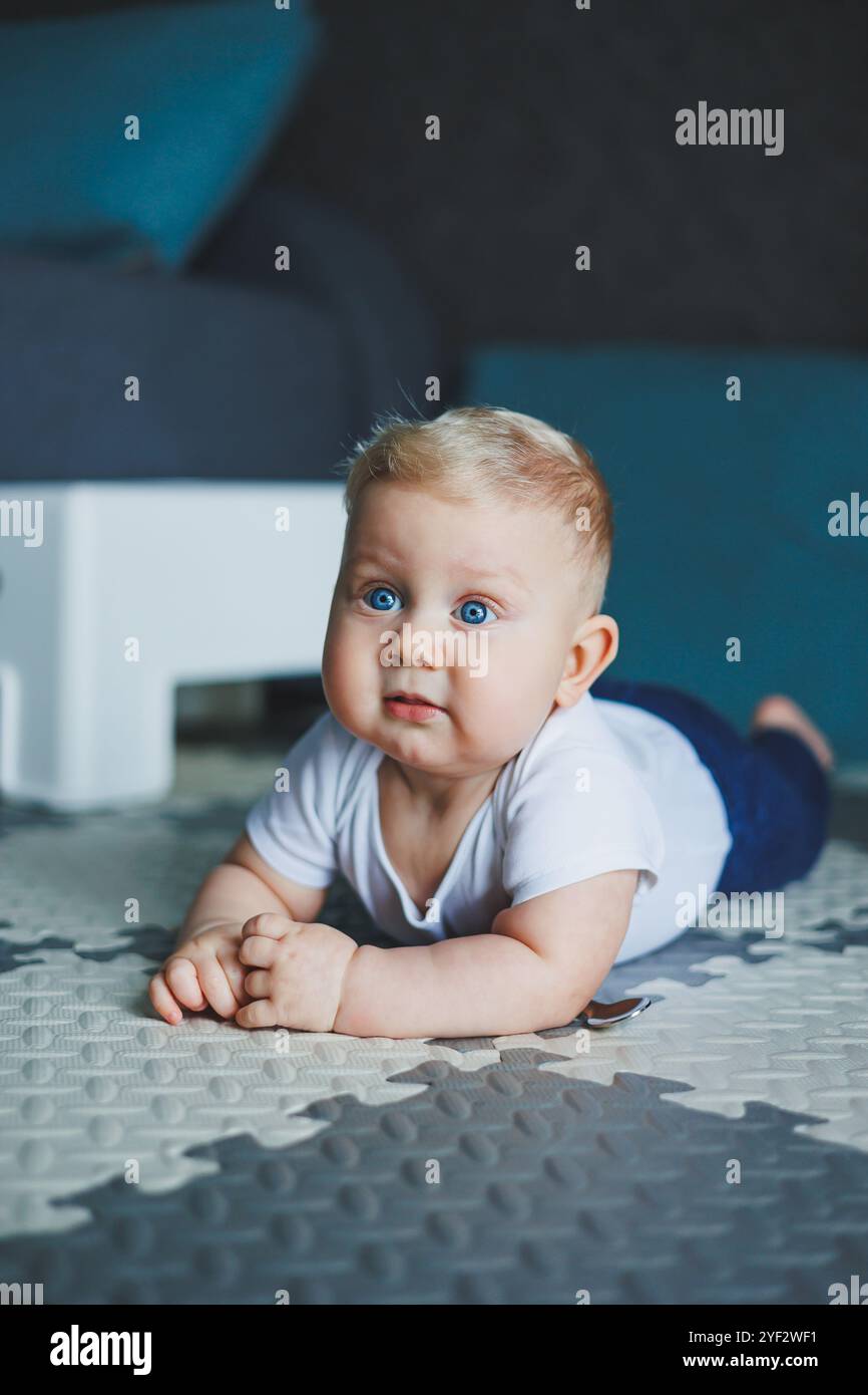 Ein blauäugiger Junge in einem weißen Body und einer blauen Hose lernt zu sitzen. Selbstentwicklung des Kindes. Das Baby sitzt auf dem Boden und lächelt Stockfoto