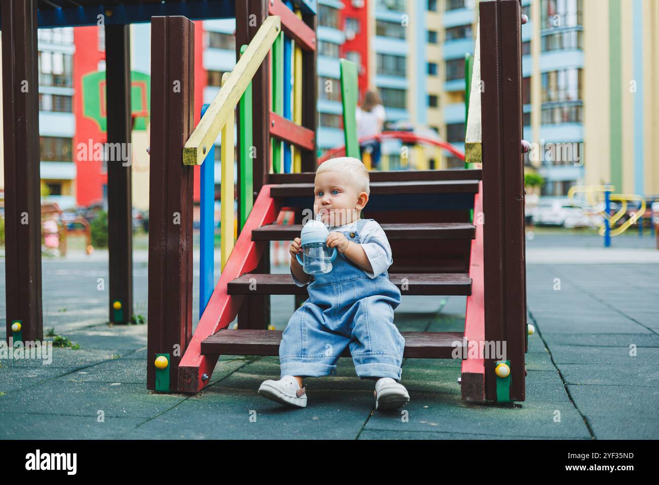 Ein 1-jähriger Junge in Denim Latzhose trinkt Wasser aus einer Flasche auf der Straße. Overall für Kinder in Übergröße. Stilvoller kleiner Junge auf dem Spielplatz. Stockfoto