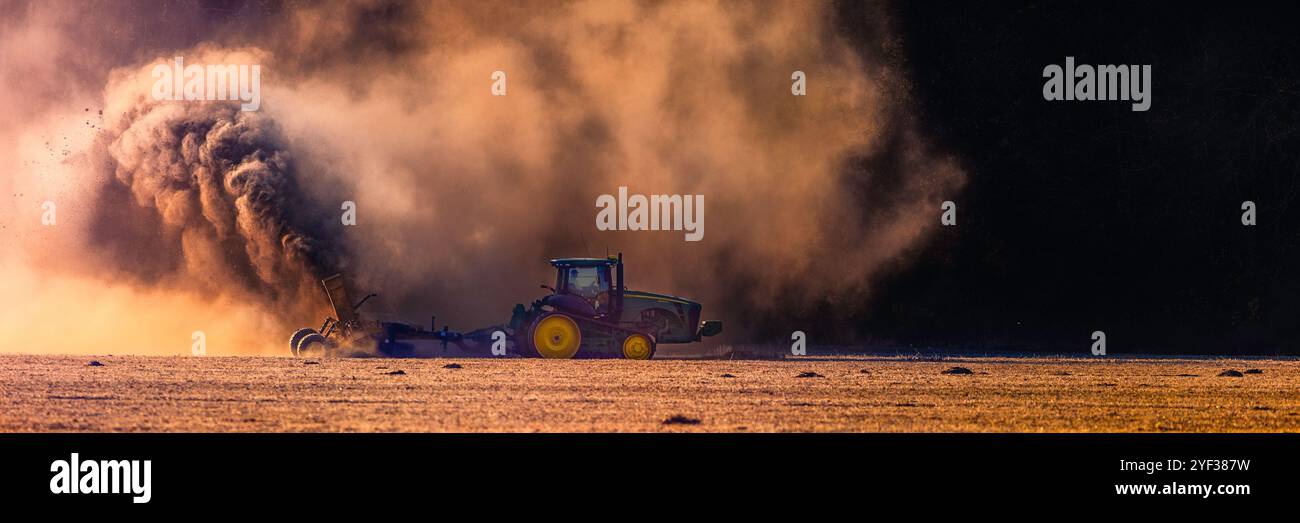 Ein großer Raupentraktor zieht ein Grabenbaugerät in Richtung dunkler Waldpartie. Viele dramatische Staubwolken werden vom Traktor aufgeschleudert Stockfoto