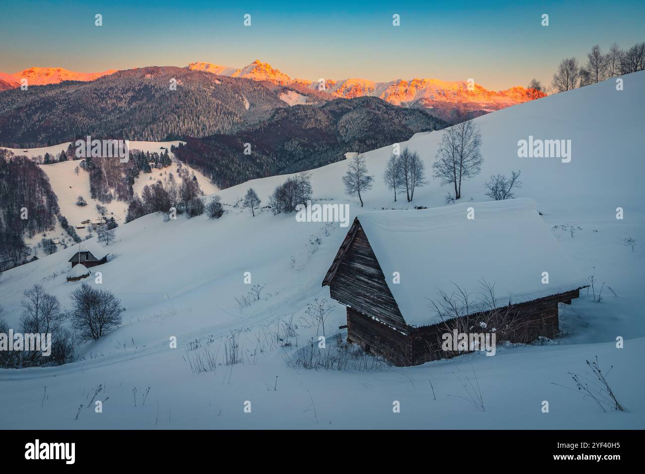 Atemberaubende Winterlandschaft mit schneebedeckter Holzhütte am Hang und spektakulären hohen Bergen im Hintergrund bei Sonnenuntergang, Bucegi Mountains, Carpathi Stockfoto