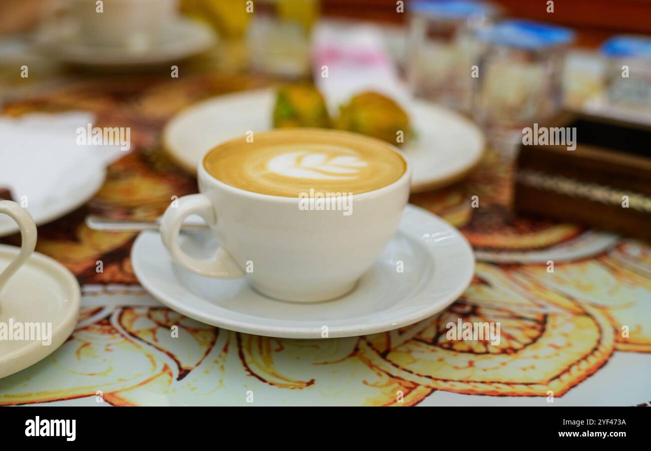 Eine warme Tasse Latte mit aufwändiger Kunst auf einem bezaubernden Tisch neben süßem Gebäck und lebendigem Obst. Stockfoto