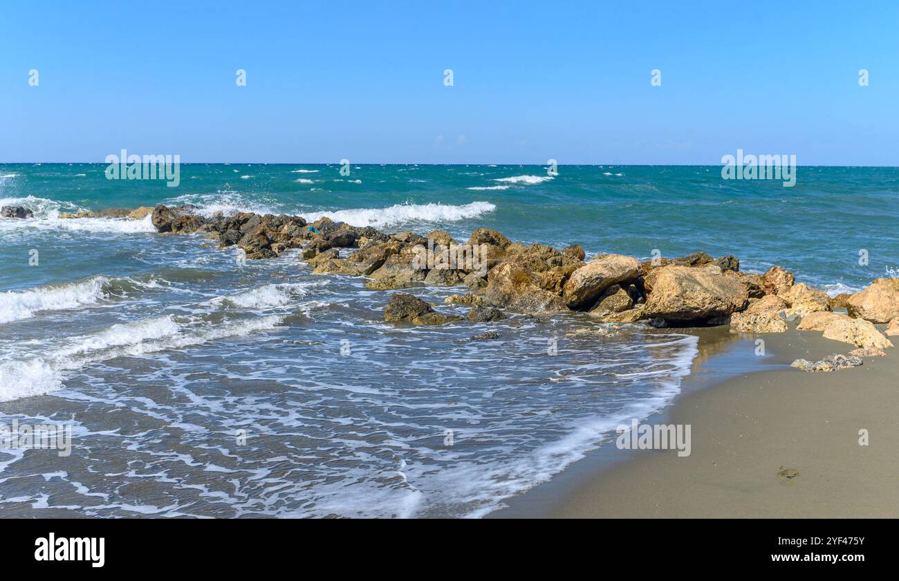 Die Sonne scheint hell, während die Wellen sanft auf die von der Sonne beschienenen Felsen entlang der ruhigen Strandküste prallen. Stockfoto