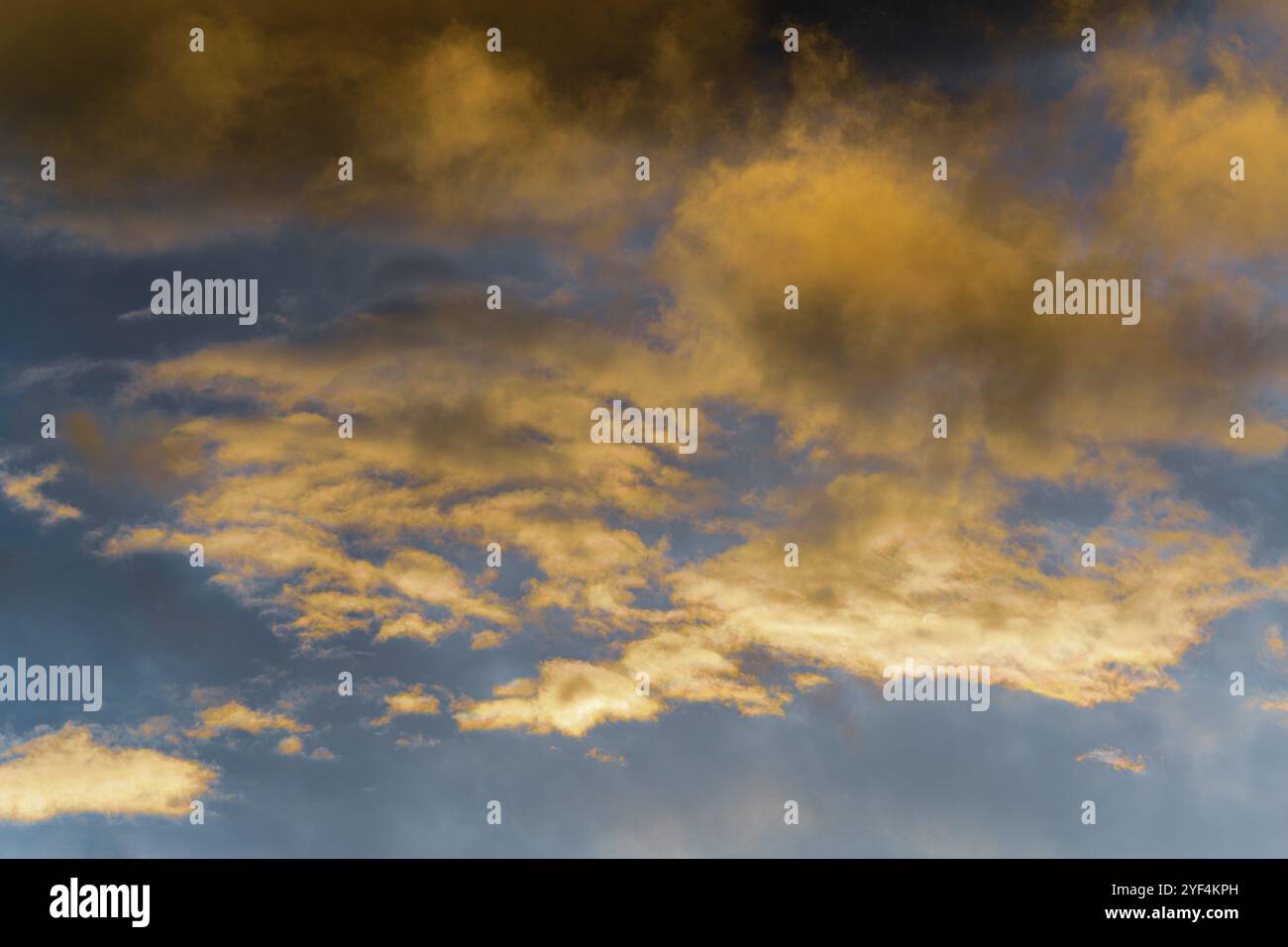 Goldene, flauschige Wolken, beleuchtet von verschwindenden Strahlen bei Sonnenuntergang und dunkle Gewitterwolken, die über den sonnigen blauen Himmel schweben, um das Wetter der Jahreszeit zu ändern. Stunni Stockfoto