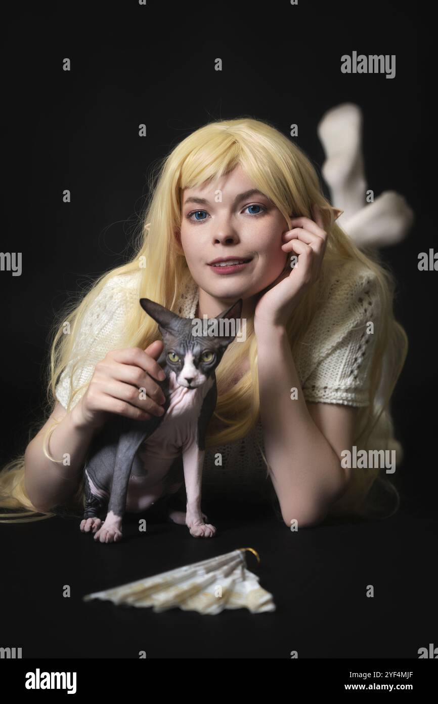 Junge weibliche Elfenkospärin in weißem Strickkleid, mit langen blonden Haaren, die auf dem Bauch auf schwarzem Hintergrund liegen, lächeln und in die Kamera schauen. Sph Stockfoto