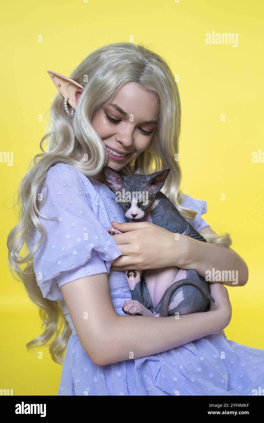 Junge Cosplay-Elfe in blauem Kleid lächelt glücklich, umarmt sanft Sphinx-Kätzchen an ihre Brust und sieht hinunter. Elf hat wunderschöne blonde Locken Stockfoto