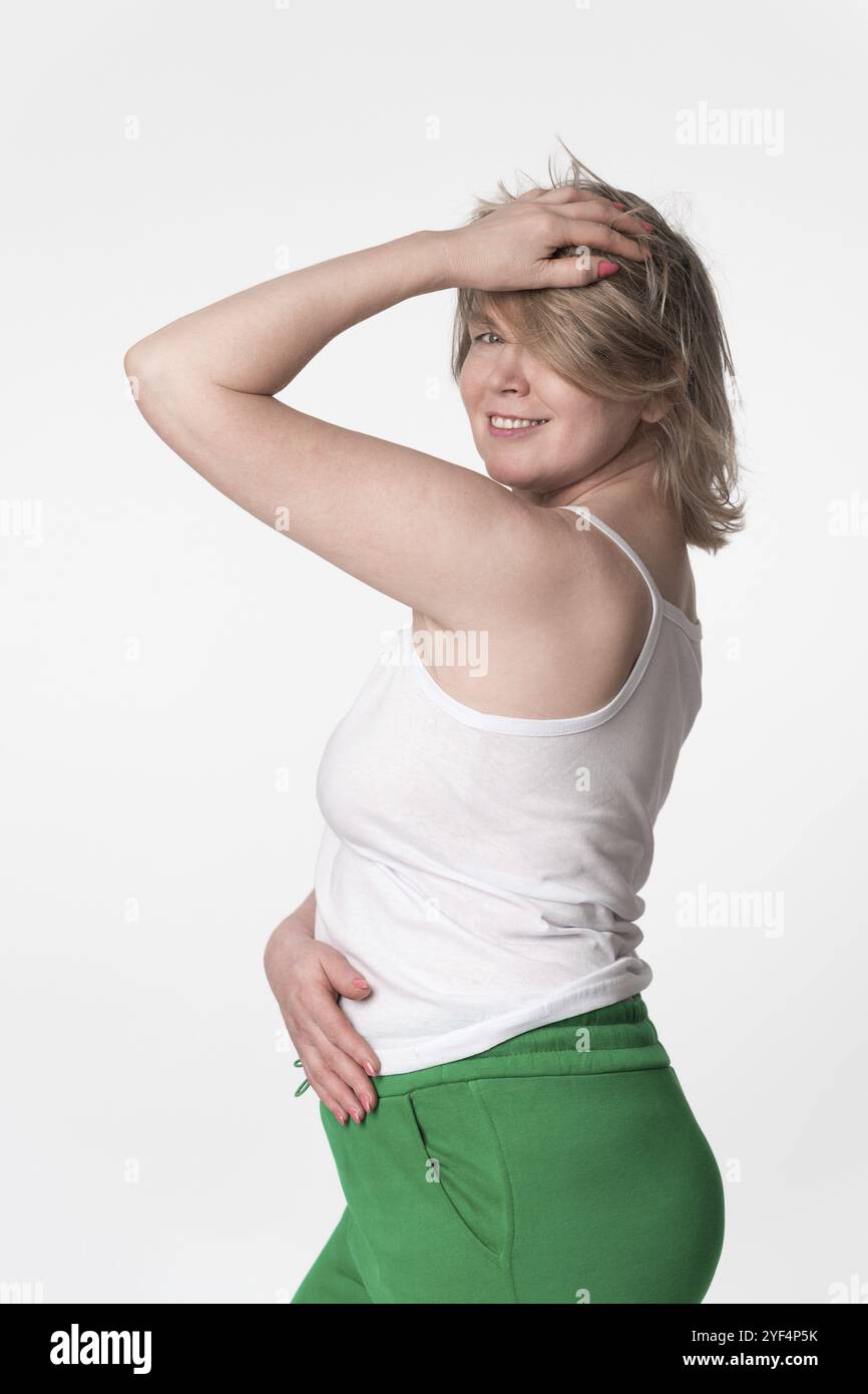 Das Porträt einer glücklichen plumpen Frau lächelt und blickt über ihre Schulter in die Kamera, posiert mit einer Hand auf dem Kopf und Hand auf dem Bauch. Sie trägt Stockfoto