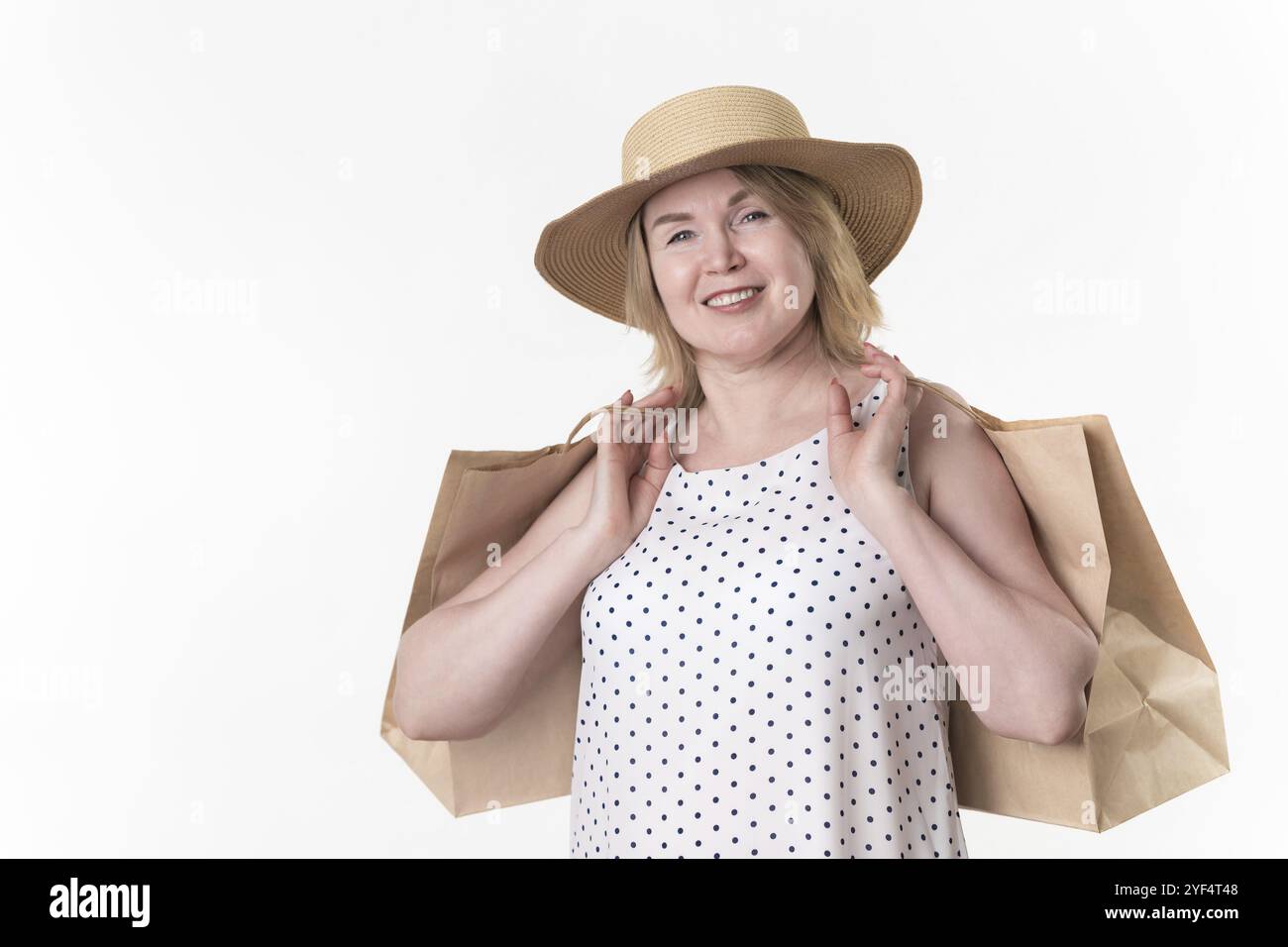 Fröhliche blonde Frau, die Papierhandwerktaschen in der Hand hält. Blonde Weibchen 49 Jahre alt in weißem Kleid mit schwarzen Polka Dots und Stockfoto