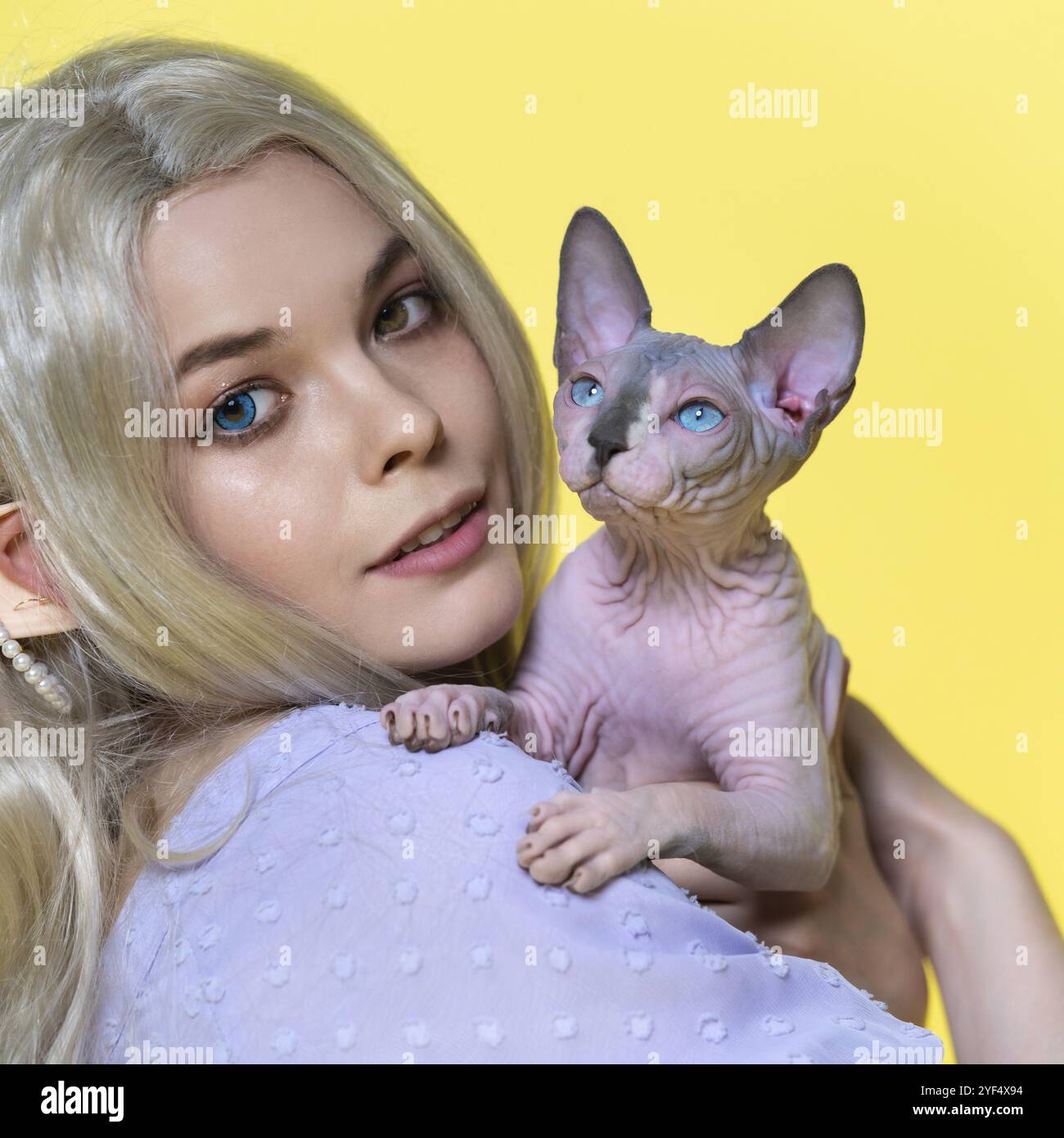 Porträt eines jungen weiblichen Cosplay-Elfs, der Sphinx-Kätzchen in der Hand hält, lächelt und mit Augen in verschiedenen Farben in die Kamera blickt. Elf hat blonde Locken Stockfoto