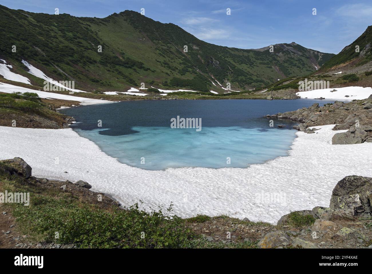 Traumhaft schöne Sommerberglandschaft der Kamchatka Halbinsel: Blauer See, Schnee und Eis entlang der Ufer des Bergsees, grüner Wald auf Hügeln Stockfoto