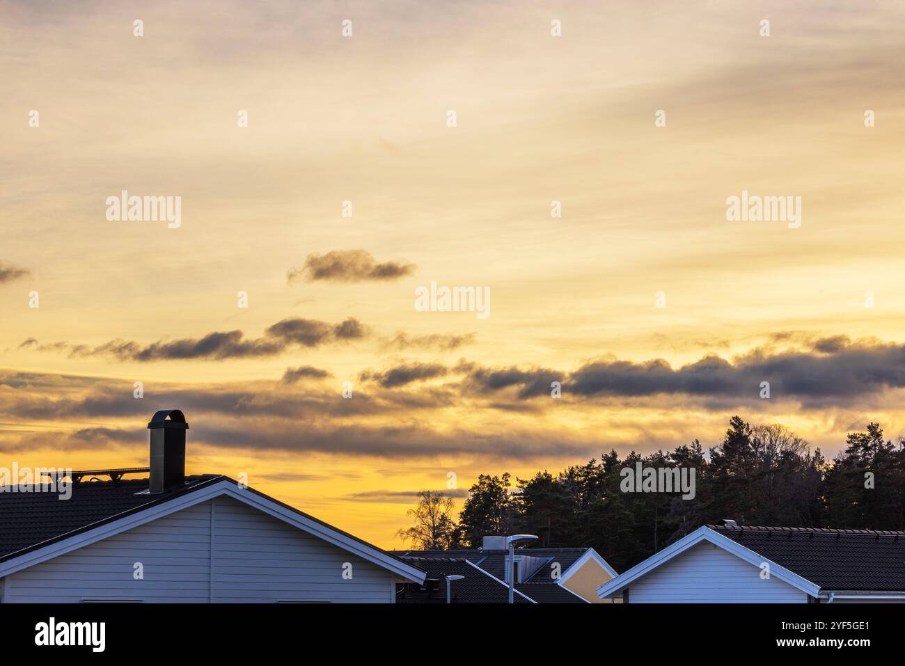 Silhouetten von Dächern und Bäumen vor dem goldenen Sonnenuntergangshimmel mit verstreuten Wolken. Stockfoto