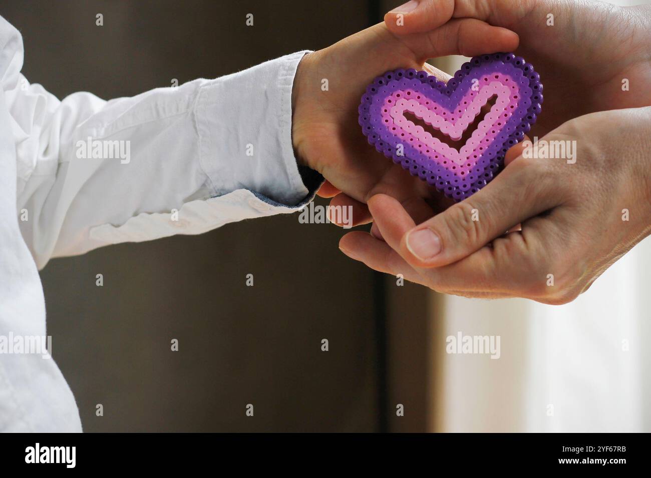 Nahaufnahme der Hand eines kaukasischen Jungen in einem weißen Hemd, der einer kaukasischen Frau ein lila rosafarbenes Herz aus Bügelperlen in die Hände gibt. Stockfoto