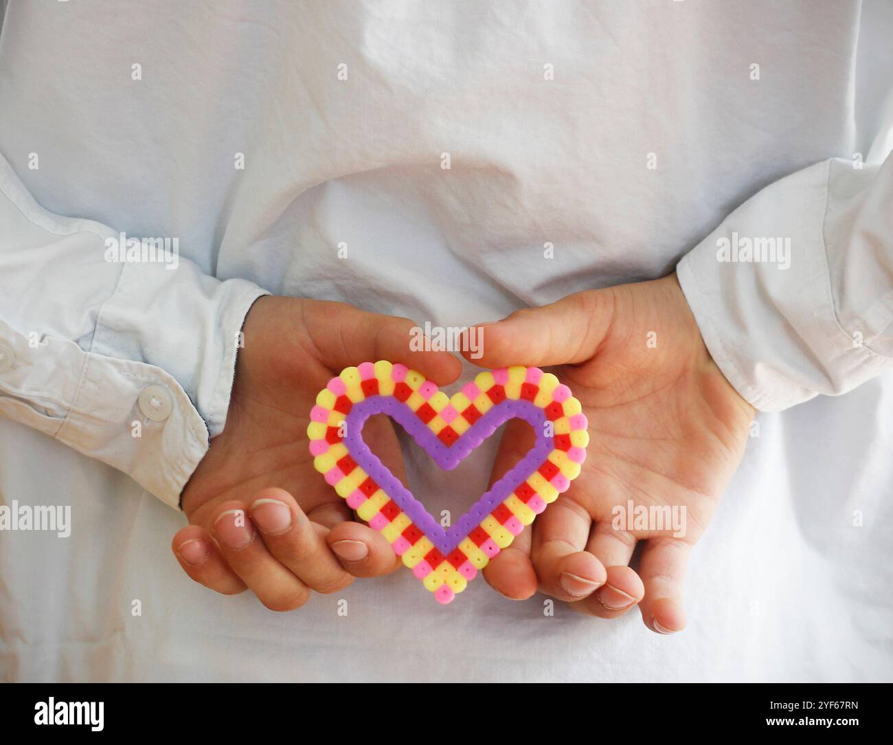 Die Hände des kaukasischen Jungen halten ein buntes Plastikherz aus Bügelperlen hinter seinem Rücken. Muttertag- und Vatertag-Konzept mit Kopierraum. Stockfoto