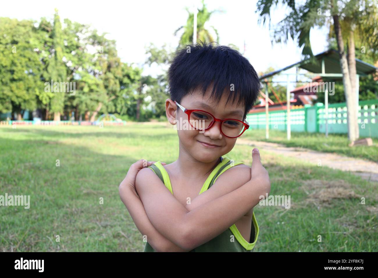 Ein charmanter Junge mit Brille lächelt fröhlich in einem sonnendurchfluteten Park, umgeben von grünem Gras und Bäumen, in dem Freude und Unschuld verkörpert werden. Stockfoto