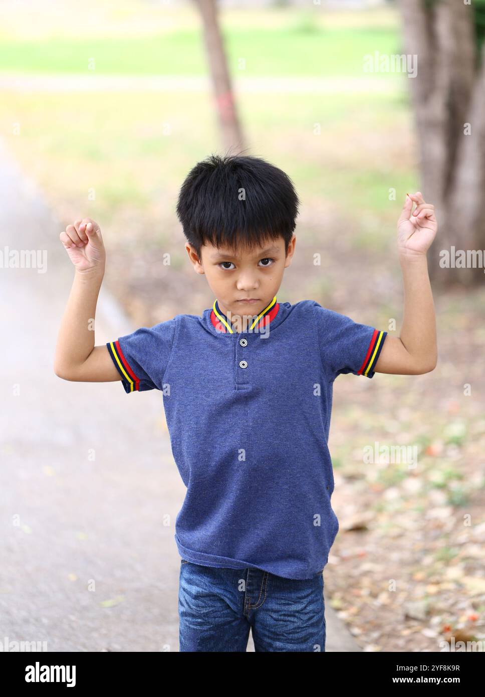 Ein selbstbewusster Junge posiert draußen und hebt seine Arme in einem Ausdruck der Freude. Die lebhafte Umgebung unterstreicht das sorgenfreie Wesen der Kindheit. Stockfoto