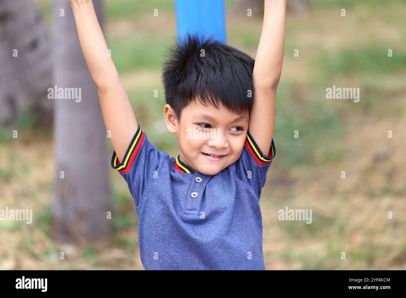 Ein fröhlicher Junge, der Spaß am Spielen im Freien hat und seine Arme vor Freude hochhebt. Sein spielerischer Ausdruck fängt das Wesen des Kinderglücks in einem lebendigen Park ein Stockfoto