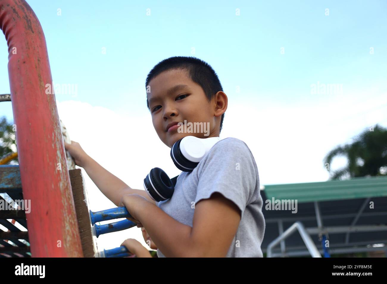 Ein fröhlicher Junge mit Kopfhörern klettert gern an einem hellen Tag auf einem Spielplatz. Sein Ausdruck verströmt Glück und Abenteuer im Freien Stockfoto