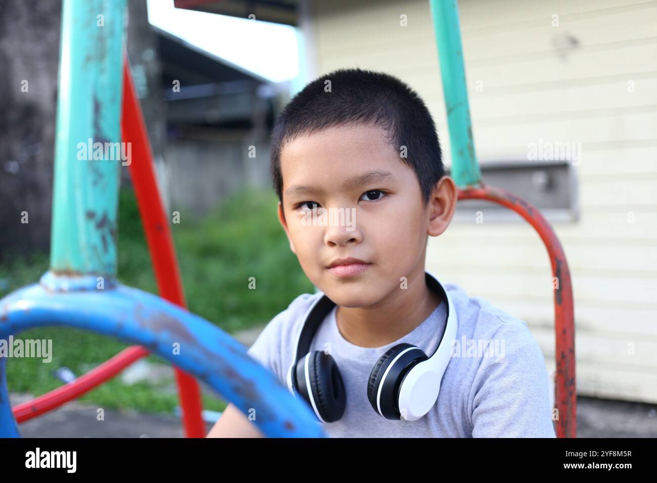 Ein kleiner Junge sitzt auf einem Spielplatz, trägt Kopfhörer, verloren in der Musik. Sein ruhiger Ausdruck verkörpert Freude und Unschuld in einem natürlichen Outdoor-Set Stockfoto