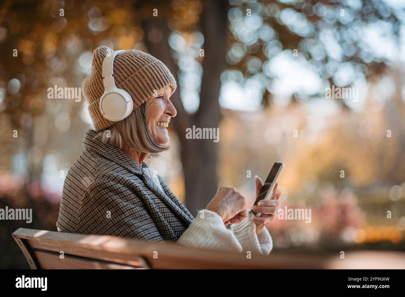 Porträt einer schönen Seniorin mit Kopfhörern im Herbstpark, Musik hören und ihr Telefon halten. Stockfoto
