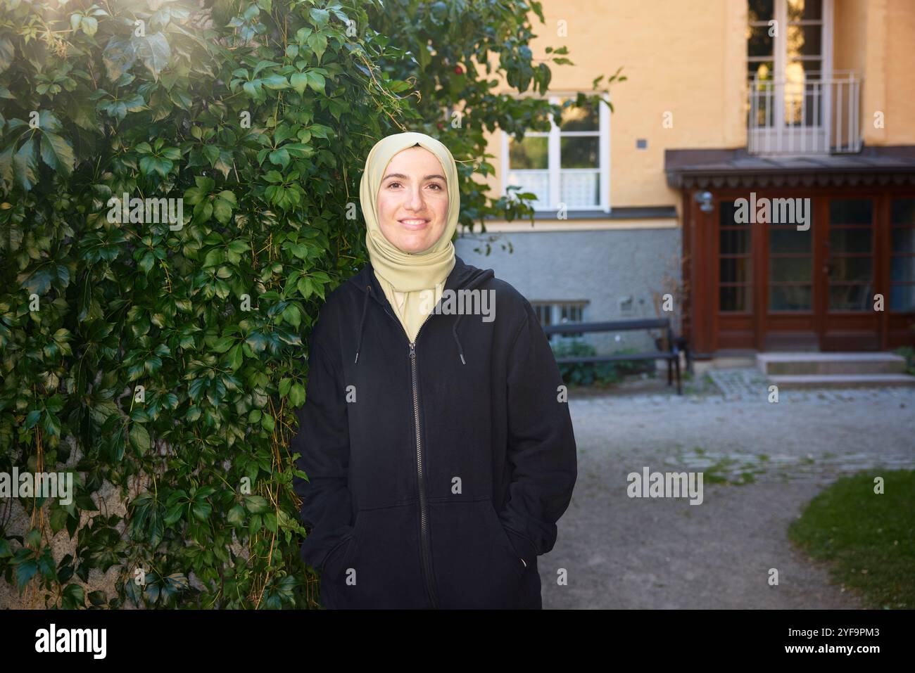 Porträt einer lächelnden, schönen Frau mit Hijab, die mit Händen in Taschen in der Nähe von Pflanzen steht Stockfoto