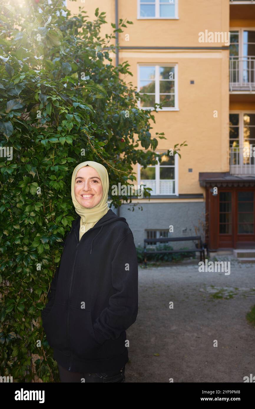 Porträt einer glücklichen, schönen Frau mit Hijab, die mit Händen in Taschen in der Nähe von Pflanzen steht Stockfoto