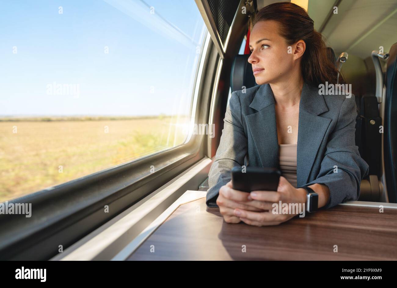 Geschäftsfrau mit Smartphone, die aus dem Fenster blickt, während sie mit dem Zug pendelt. Geschäftsreisekonzept. Stockfoto