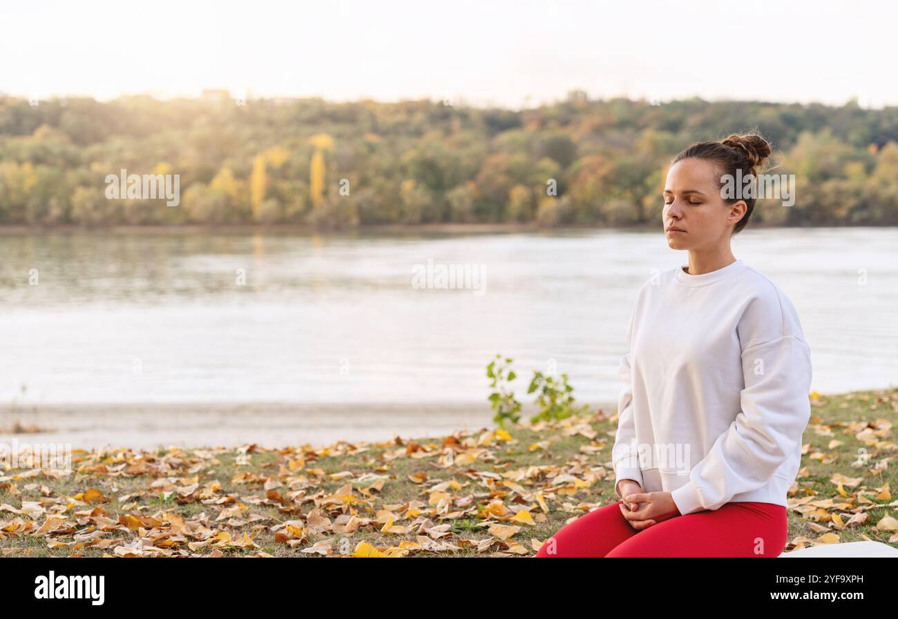 Ruhige Frau, die am Fluss im Herbst Yoga praktiziert und die Natur und das Wohlbefinden einnimmt. Stadtrelaub. Stockfoto