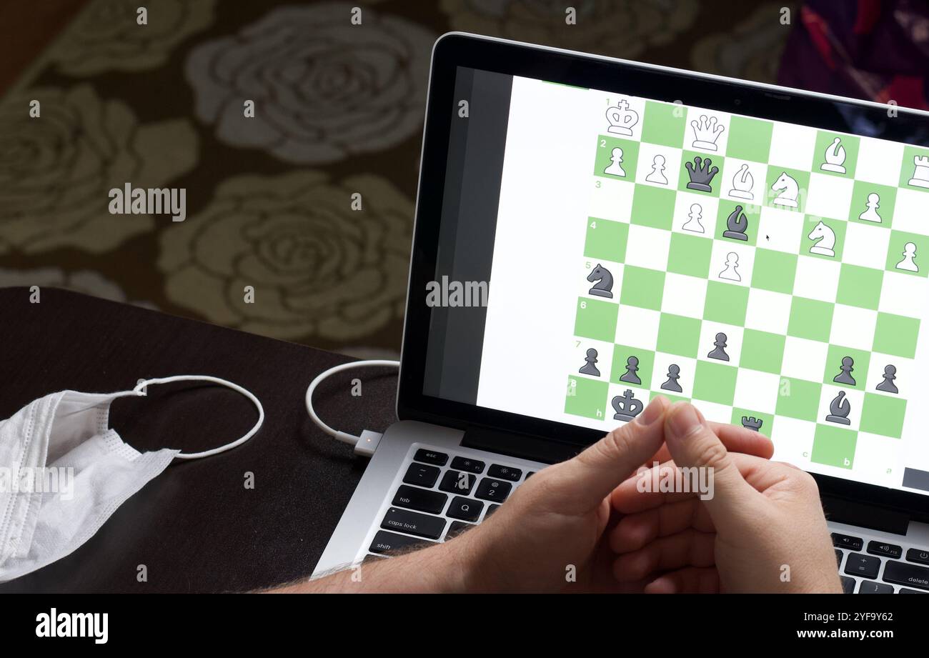 Ein Mann löst online ein Schachproblem. Schachbrett mit Figuren auf einem Laptop-Bildschirm. Freizeit in Selbstisolierung, in der Dämmerung. Internationaler Schachtag. Stockfoto
