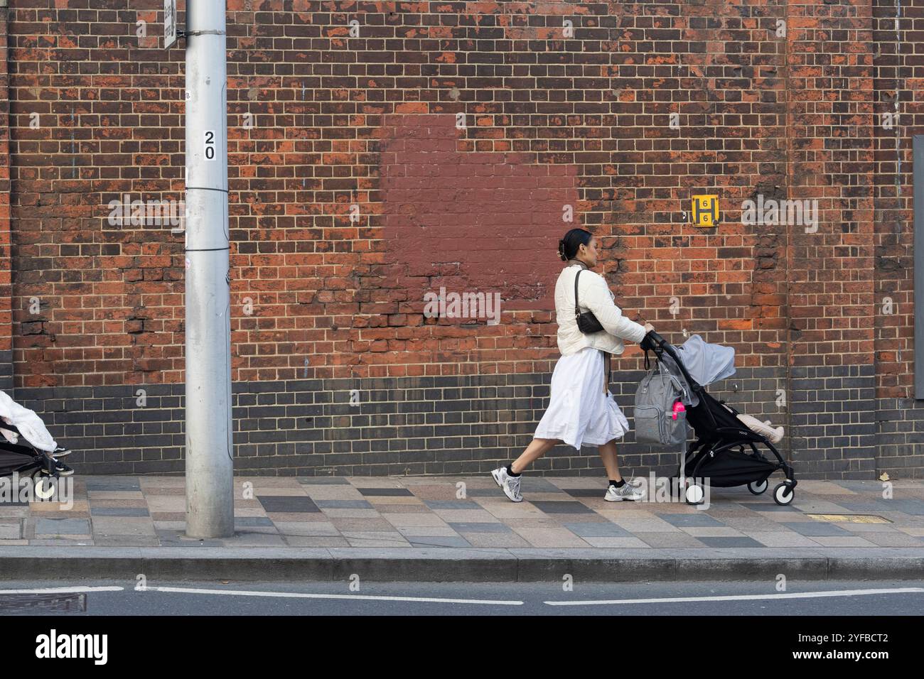 Frau, die einen Kinderwagen entlang der Straße schiebt, wahrscheinlich in der Nähe eines Gebäudes mit Adresse 20, auf einem Bürgersteig. London, Großbritannien. Stockfoto